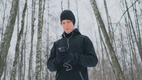 A-young-man-in-a-black-trigger-in-the-winter-is-preparing-to-jog-in-the-park-includes-running-music-on-a-smartphone-and-runs-with-a-voice-assistant-through-the-forest-doing-business-and-leading-a-healthy-lifestyle